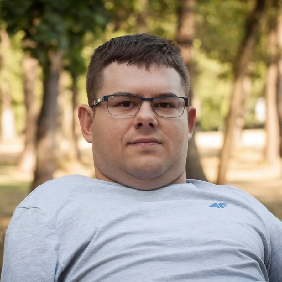 Portrait of Łukasz Socha wearing glasses and a light gray shirt, photographed outdoors against a softly blurred green background.